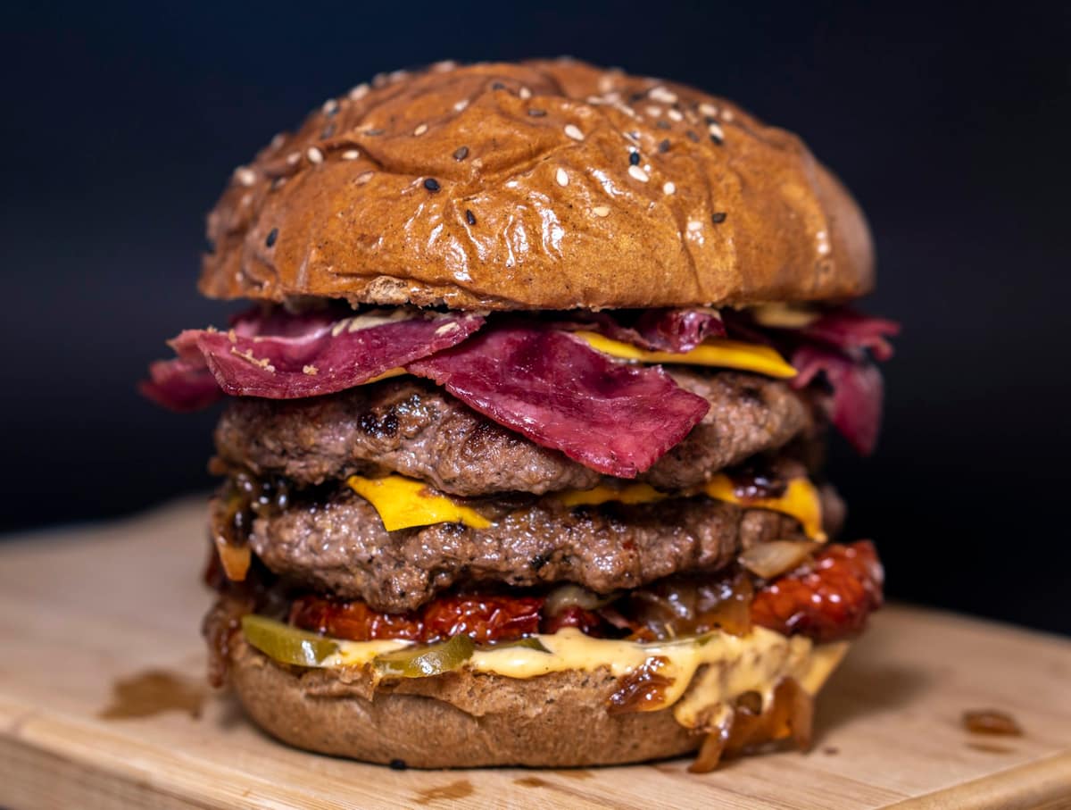 Hamburguesa apilada con fetas de carne ahumada, queso cheddar y salsa en pan de sésamo sobre tabla de madera