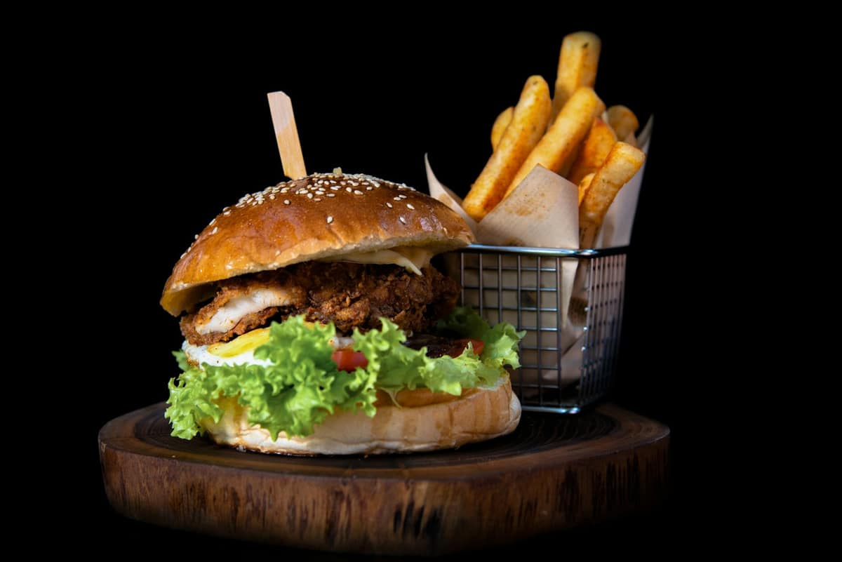 Hamburguesa crocante con lechuga junto a una cesta de papas fritas sobre tabla de madera, fondo negro