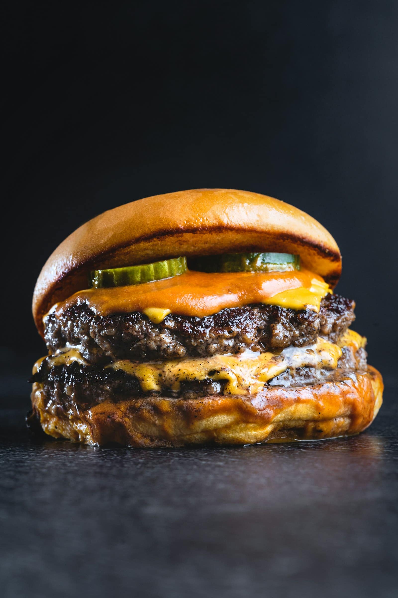 Hamburguesa doble con queso cheddar fundido, pepinillos y salsa, fotografiada de cerca sobre fondo oscuro